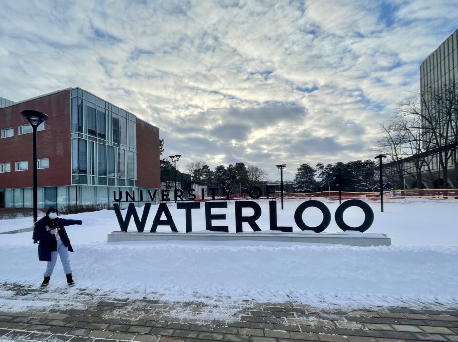 Image of Nabiha a young woman standing in her winter coat and mask to the side of the University of Waterloo sign outside on campus.