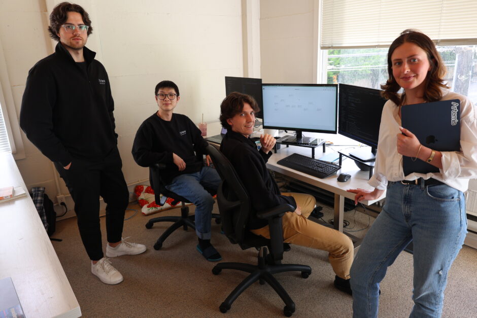 Artemis Data staff and co-founders stand in front of a desk with three computer monitors on it.