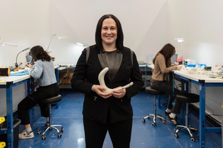 
Rachel Bartholomew, CEO and co-founder of Hyivy Health holding the Floora, the company’s Bluetooth-connected pelvic rehabilitation device. Two women are behind her sitting by workstations with their heads down working.