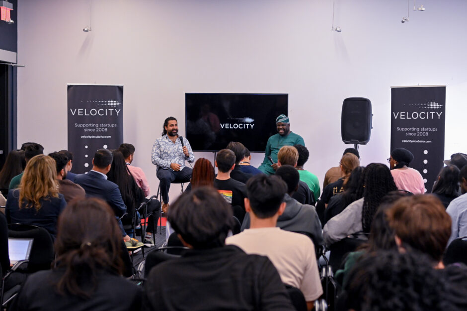 Akash Vaswani on the left and Iyinoluwa Aboyeji on the right. In between them is a screen with the Velocity logo and on either side is a banner with the Velocity logo. They are sitting in front of a group of attendees.